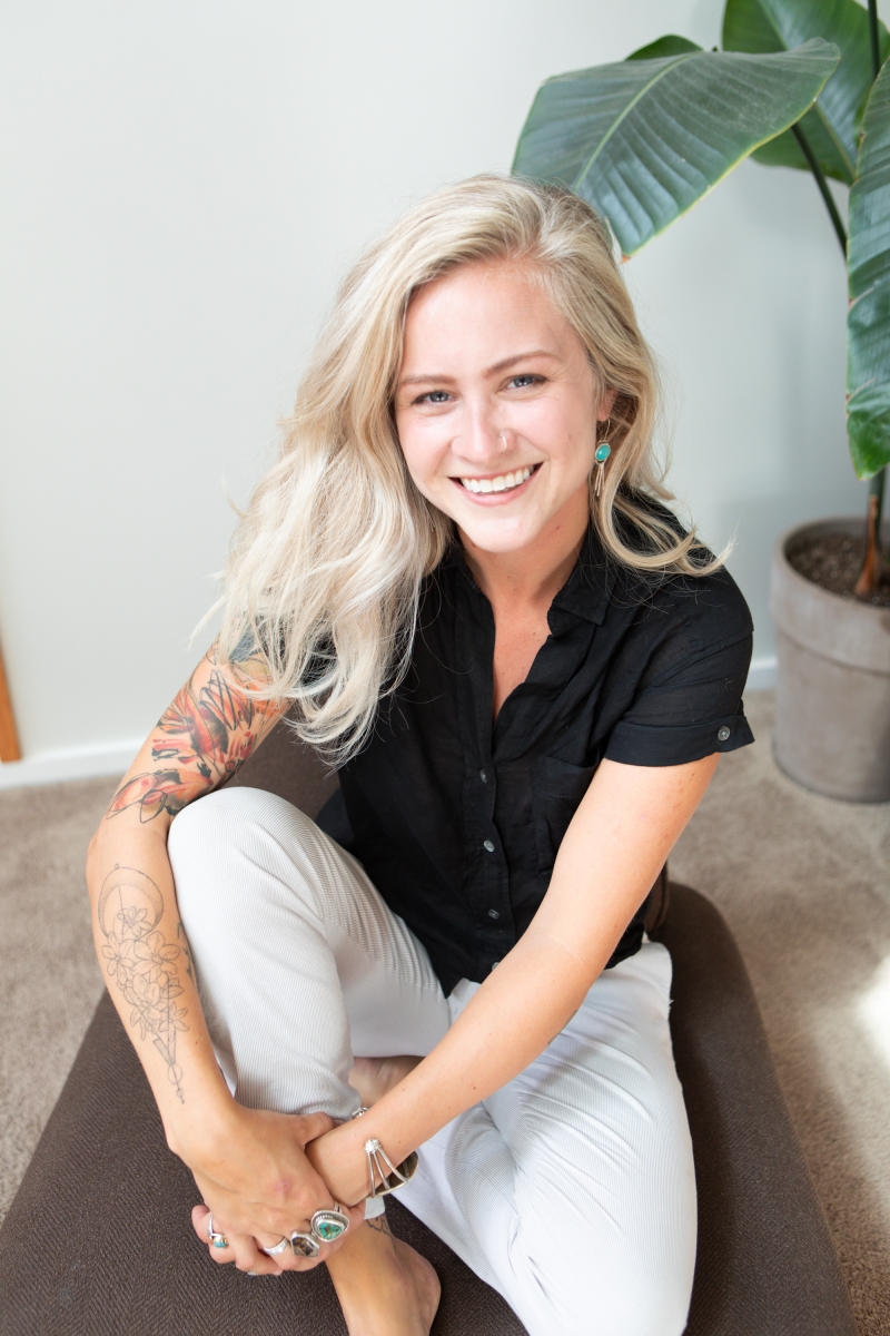 A blond girl sitting on a chair, smiling, with a green plant behind her.